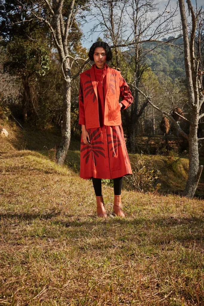 The image displays a woman wearing a rustic red dress and a quilted jacket with floral patterns. Copyright Khara Kapas.
