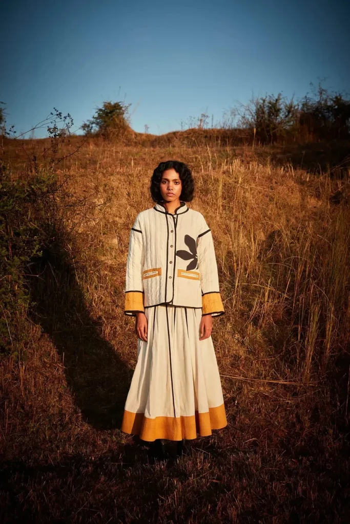 The image displays a woman wearing an off-white mul jacket and maxi skirt, detailed with an intricate floral patchwork. Copyright Khara Kapas.