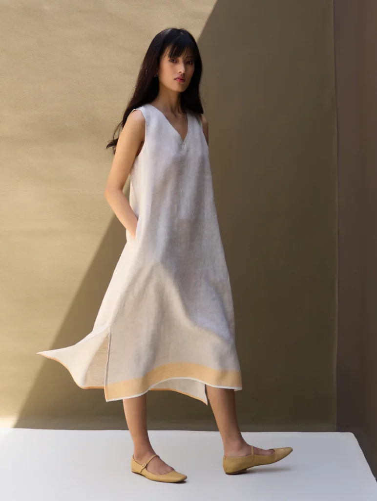 The image displays a woman wearing a handwoven, minimalist, sleeveless dress with a v-neck design in oat and orange. Copyright Manan.