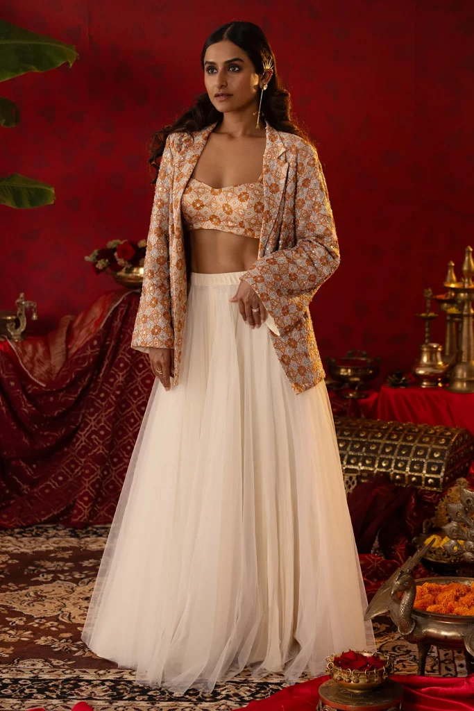 The image displays a woman wearing a beige ajrakh blazer embellished with jeweled sequins, jardosi, zari, and an off-white tulle skirt and bustier. Copyright Pooja Bagaria.
